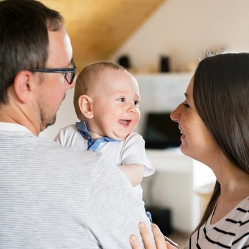Eine junge Familie zu Hause. Der Vater hält ein lachendes Baby auf dem Arm. Die Mutter und das Baby schauen sich an. 