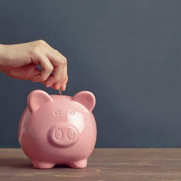 Es ist eine Hand zu sehen, die eine Münze in ein rosa Sparschwein gibt.