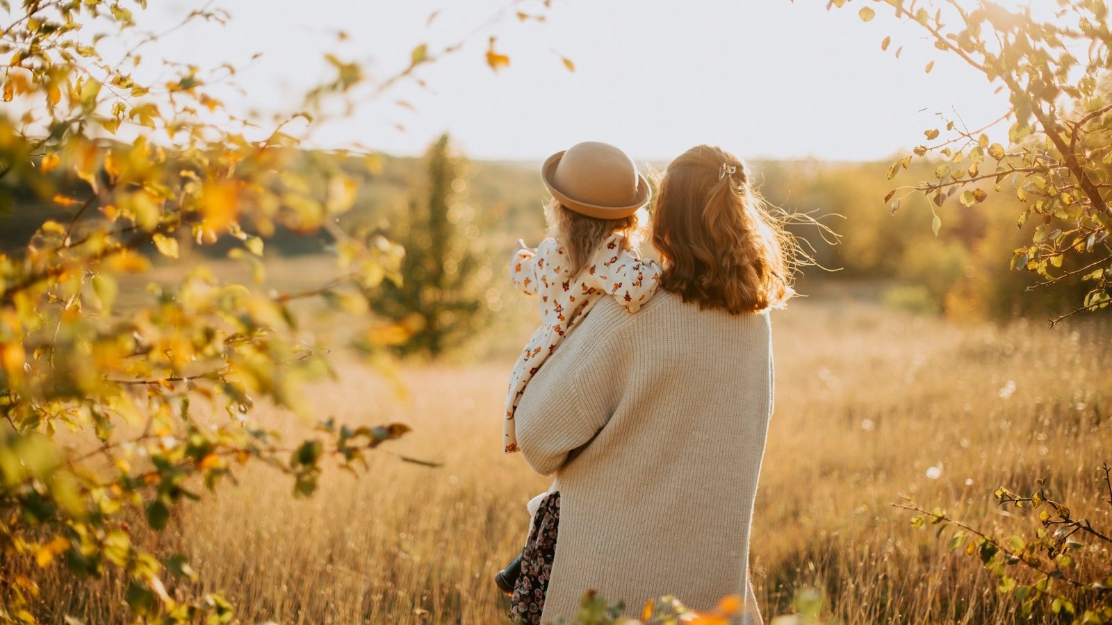 Eine Frau steht mit einem kleinen Kind auf dem Arm in einer Wiese, die in goldener Herbstfarbe strahlt.