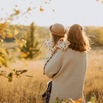 Eine Frau steht mit einem kleinen Kind auf dem Arm in einer Wiese, die in goldener Herbstfarbe strahlt.