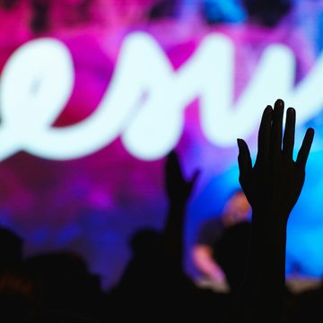 Menschen halten ihre Hände in die Höhe, im Hintergrund leuchtet ein Schild auf dem "Jesus" steht