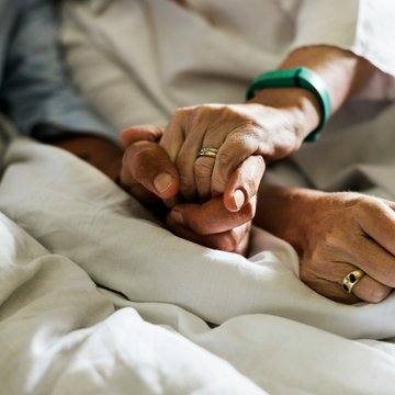 Die Hände von zwei älteren Menschen halten sich gegenseitig. Die beiden Menschen sitzen halb auf in einem Bett.