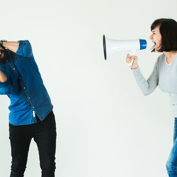 Eine Frau und ein Mann stehen nebeneinander, die Frau hält ein Megaphon vor dem Mund und hält es in die Richtung des Mannes. Der Mann hält sich die Ohren zu.