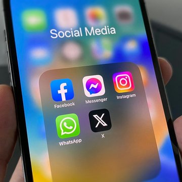 Ein Smartphone wird in einer Hand gehalten, der Bildschirm zeigt einen Ordner der mit "Social Media" überschrieben ist und die App-Symbolde von Facebook, Facebook Messanger, Instagram, WhatsApp und X (ehemals Twitter) zeigt.