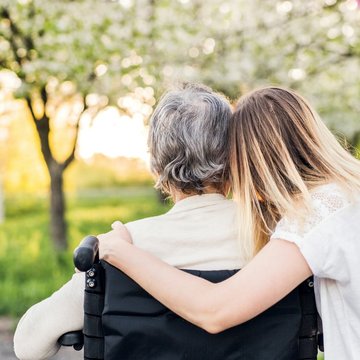 Eine ältere Frau sitzt im Rollstuhl. Eine junge Frau hat ihren Arm um sie gelegt. Im Hintergrund sind Bäume und eine Wiese zu sehen. Die beiden Frauen sind von hinten zu sehen.