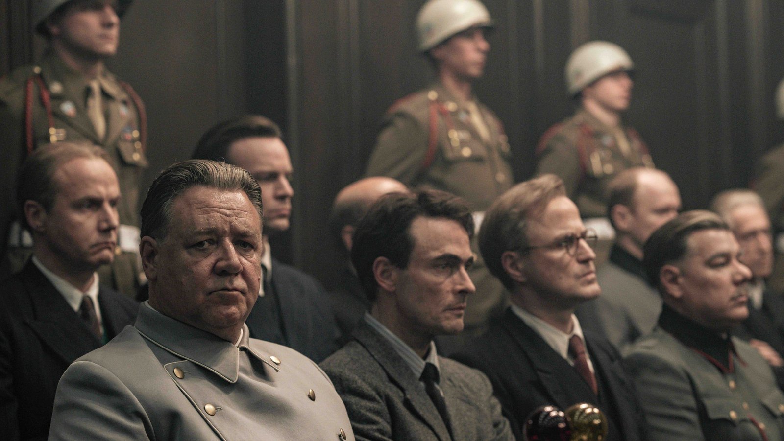 Das Bild zeigt einen Ausschnitt aus dem Film "Nürnberg": Hermann Göring (links), gespielt von Russell Crowe, sitzt mit anderen Männern auf der Anklagebank in Nürnberg. Im Hintergrund sind Wachen in Uniform zu sehen.