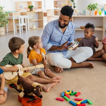Ein Mann sitzt zwischen kleinen Kindern auf dem Boden mit Spielsachen um alle herum. Eines der Kinder hat eine kleine Gitarre in der Hand.