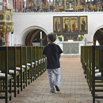 Ein Kind läuft durch den Mittelgang einer Kirche in Richtung Altar. Rechts und links sind leere Stühle aufgestellt.