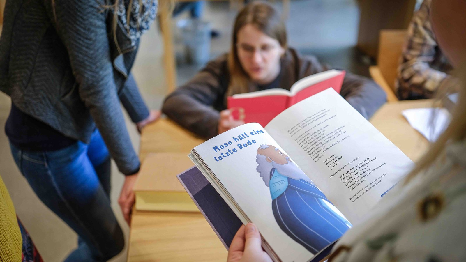 Frauen sitzen und stehen um einen Tisch herum und lesen Bücher. Über die Schulter der vorderen Frau sieht man ein aufgeschlagenes Buch auf dem "Mose hält eine Rede" steht.