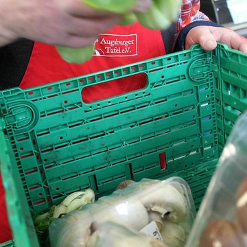 Das Bild zeigt eine Person, die aus einer grünen Kiste einen Salatkopf nimmt. In der Kiste sind noch verpackte Pilze zu sehen.