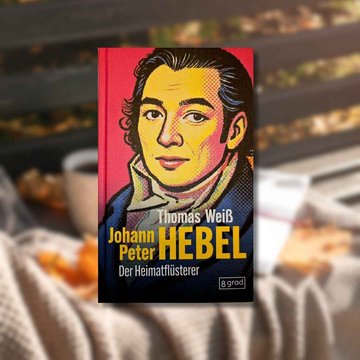 Buchtipp: „Johann Peter Hebel – Der Heimatflüsterer“ im Vordergrund. Im Hintergrund ist eine Bank zu sehen, auf der herbstliche Blätter liegen, sowie eine Tasse und ein Buch.