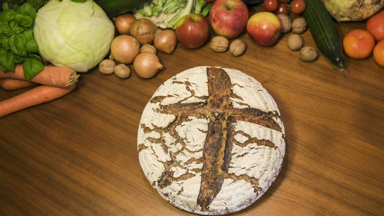 Ein Laib Brot mit einem Kreuz und Obst und Gemüse im Hintergrund liegen auf einem Tisch 