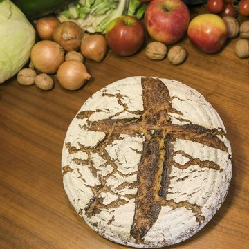 Ein Laib Brot mit einem Kreuz und Obst und Gemüse im Hintergrund liegen auf einem Tisch 