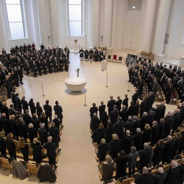 Das Foto zeigt eine große Menge an schwarz gekleideten Menschen in der St. Hedwigs-Kathedrale in Berlin. Sie stehen vor ihren Stühlen, die im Kreis um eine Halbkugel aus Stein (Taufstein) aufgestellt sind.