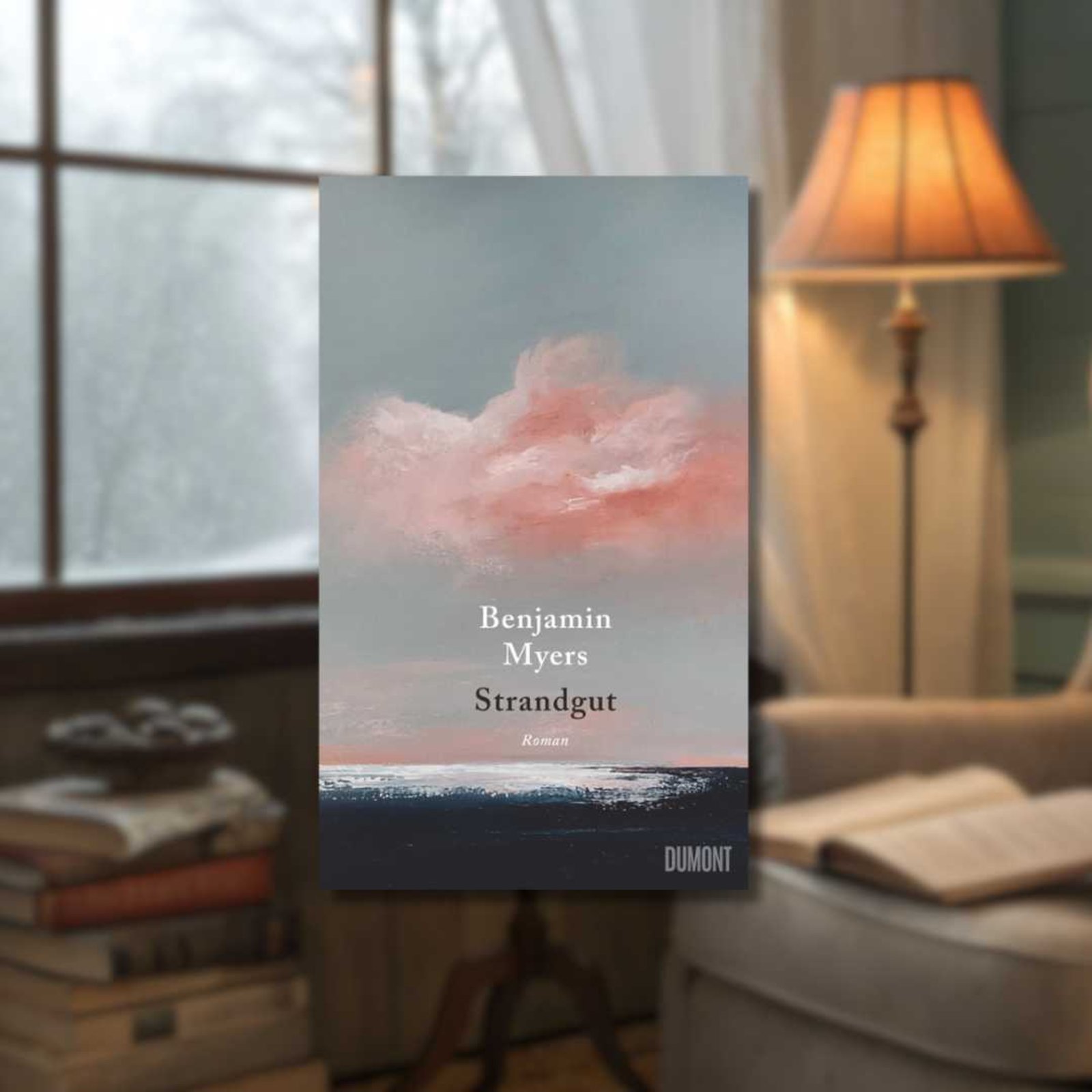 Buchcover „Benjamin Myers: Strandgut” im Vordergrund, im Hintergrund ein gemütlich eingerichtetes Wohnzimmer mit einer angeschalteten Stehlampe, Bücherstapel auf dem Boden und ein aufgeschlagenes Buch auf der Sitzfläche eines Sessels.