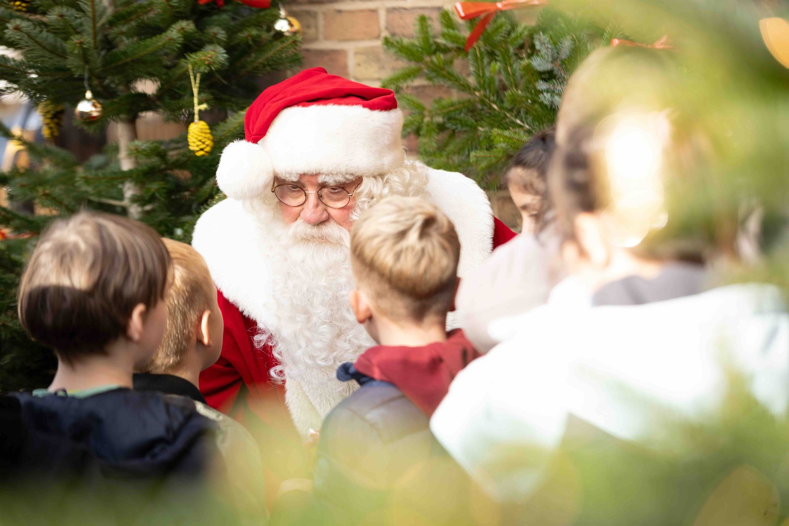 Ein Mann in einem Weihnachtsmannkostüm (rot-weiße Mütze, langer weißer Bart und roter Mantel) sitzt vor einem geschmückten Christbaum. Vor dem Weihnachtsmann stehen Kinder und schauen ihn an.