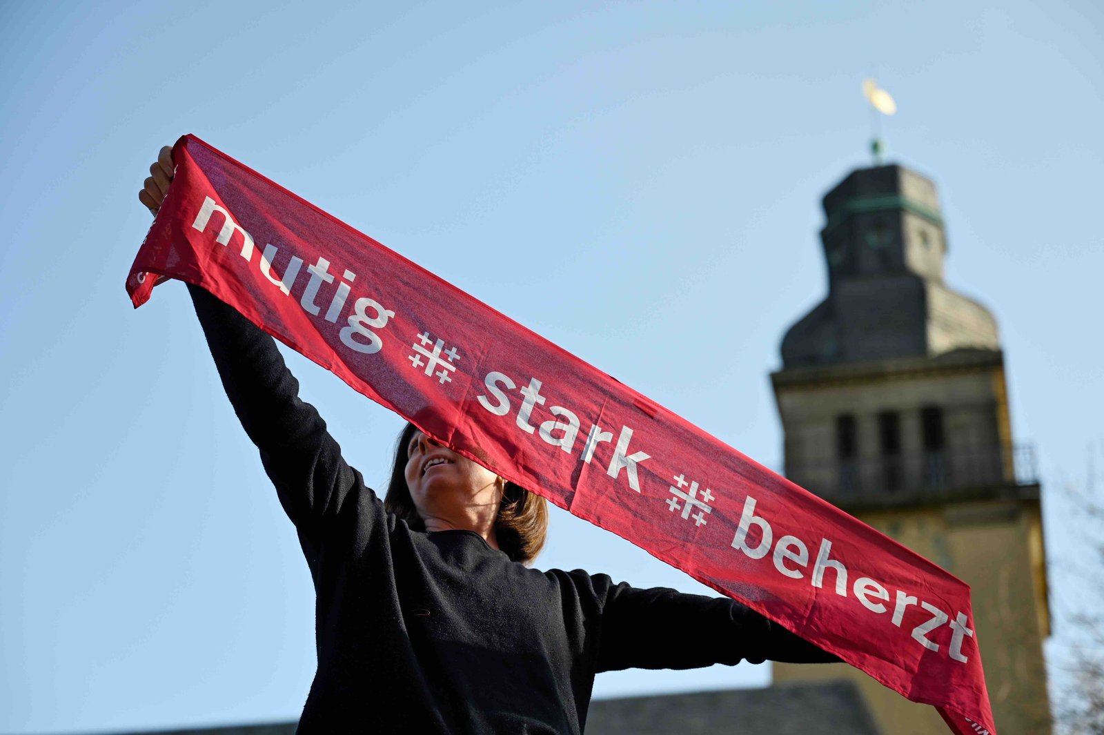 Eine Frau hält einen Banner hoch auf dem steht: „mutig + stark + beherzt”
