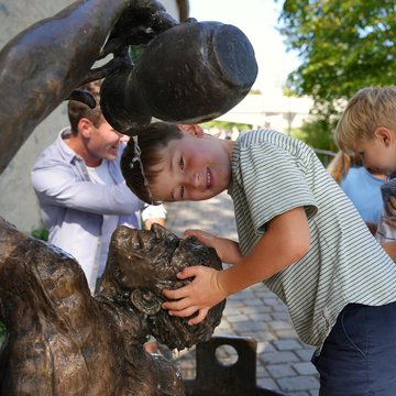 Ein Kind hält den Kopf zwischen eine Statue, die gleichzeitig ein Brunnen ist.