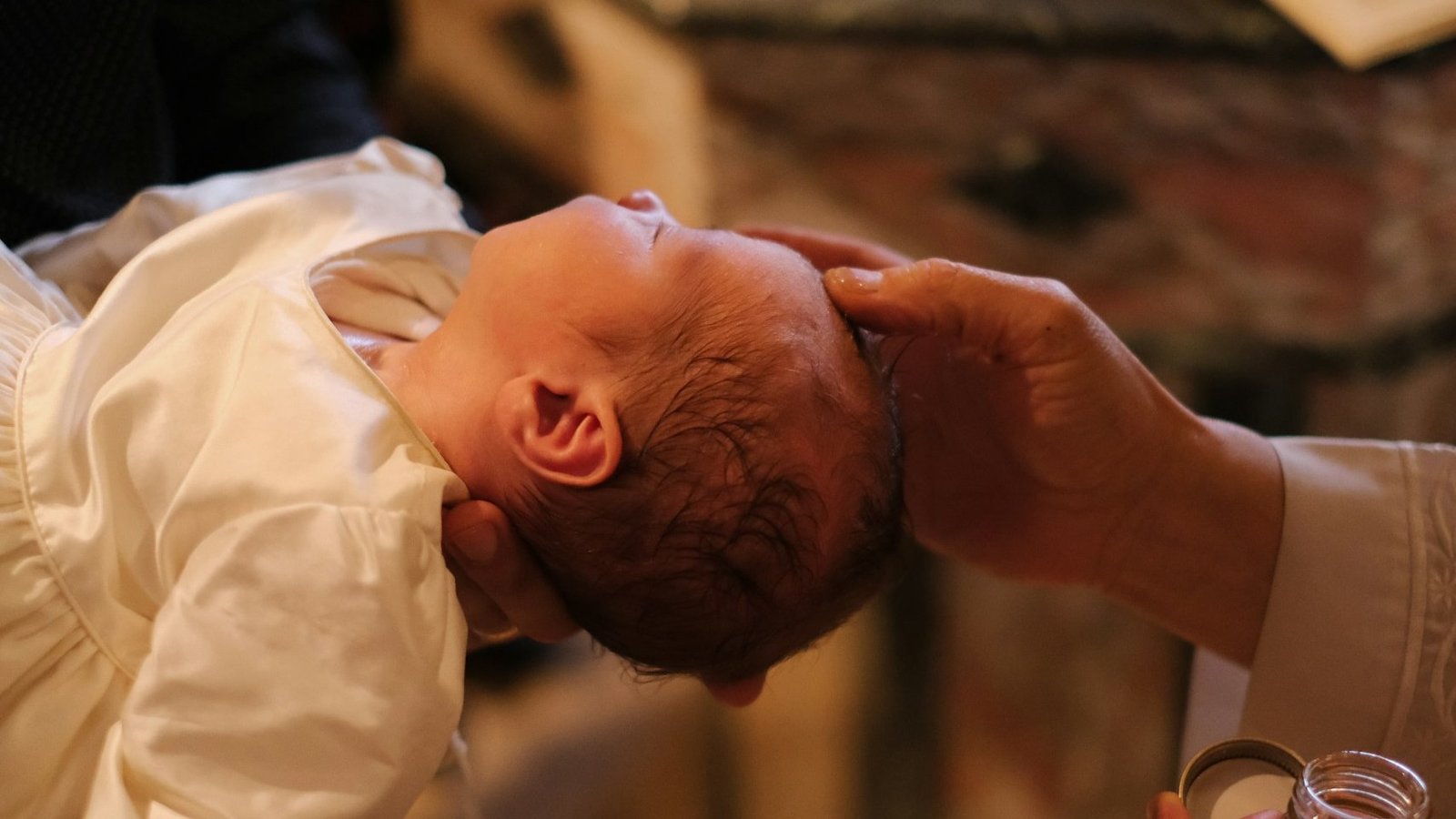 Ein Baby bei seiner Taufe. Eine Person hält das Baby auf den Armen, ein Pfarrer hält seine Hand an den Kopf des Babys