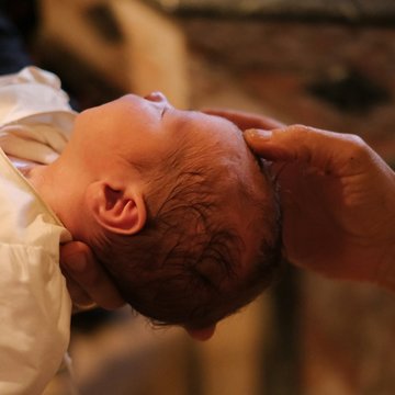 Ein Baby bei seiner Taufe. Eine Person hält das Baby auf den Armen, ein Pfarrer hält seine Hand an den Kopf des Babys