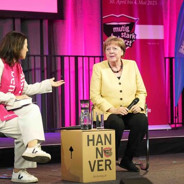 Links sitzt Anja Siegesmund, rechts neben ihr Angela Merkel auf einer Bühne. Im Hintergrund stehen Banner mit dem Logo des 39. Evangelischen Kirchentags in Hannover