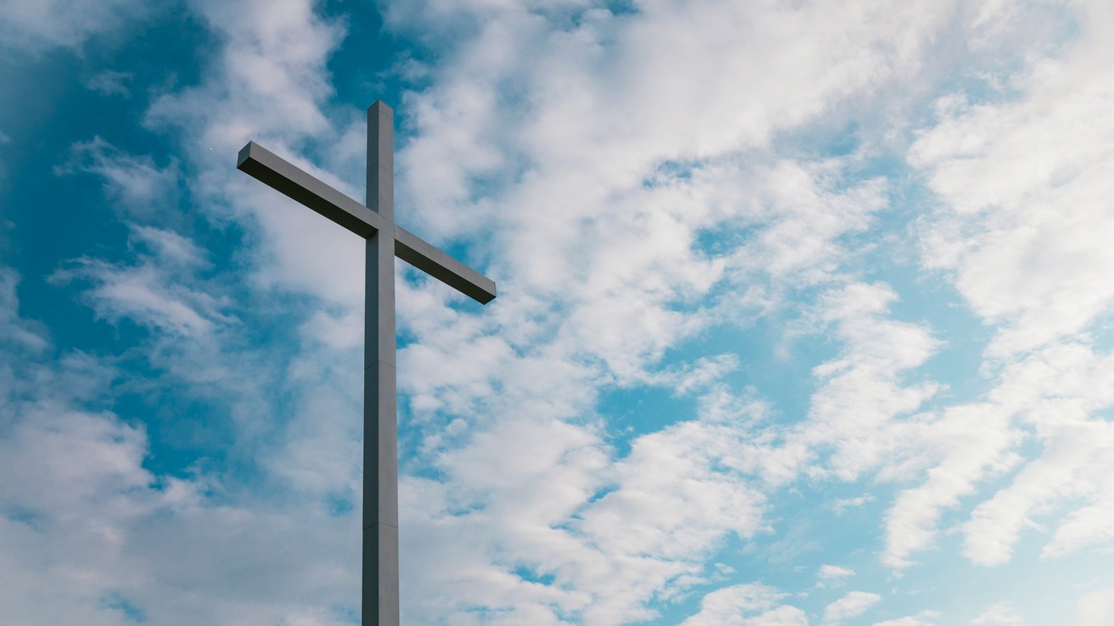 Ein Kreuz aus grauem Material ist von unten fotografiert gegen den Himmel, an dem weiße Wolken zu sehen sind.
