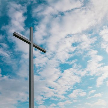 Ein Kreuz aus grauem Material ist von unten fotografiert gegen den Himmel, an dem weiße Wolken zu sehen sind.