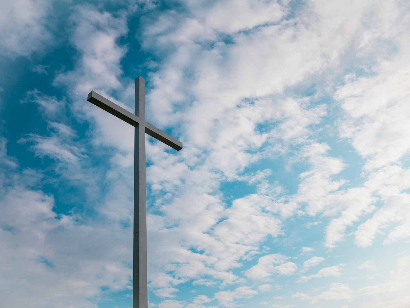 Ein Kreuz aus grauem Material ist von unten fotografiert gegen den Himmel, an dem weiße Wolken zu sehen sind.