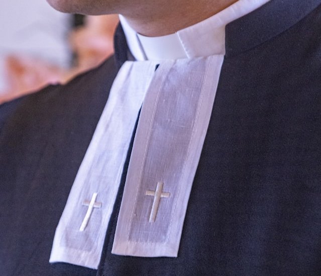 Nahaufnahme eines Mannes in einem schwarzen Talar mit weißem Beffchen auf dem zwei Kreuze eingestickt sind. 