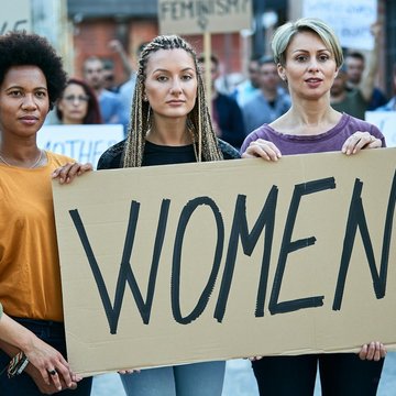 Fünf Frauen stehen nebeneinander und halten ein Papp-Schild in der Hand auf dem "Women" (Frauen) steht. Die Frauen haben einen ernsten Gesichtsausdruck.