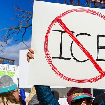 Menschen stehen nebeneinander. Eine Person hält ein Schild hoch auf dem "ICE" steht. Um die Buchstaben ist ein roter Kreis gemalt und ein roter Strich geht innerhalb des Kreises schräg über die Buchstaben.