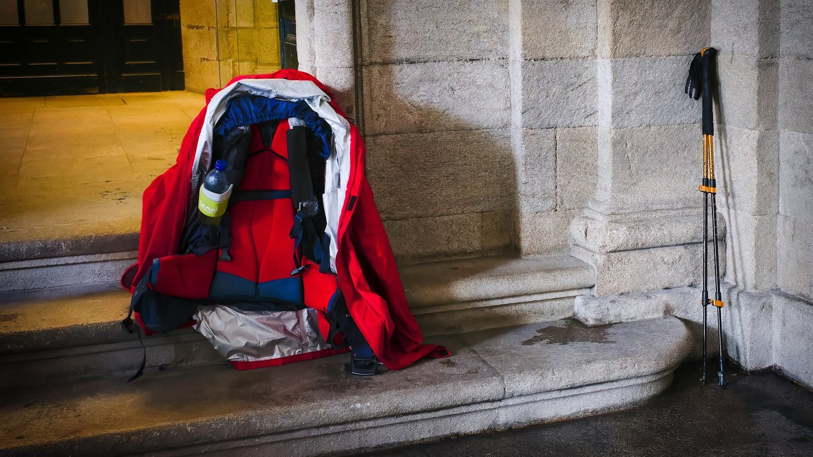 Ein Rucksack, der auf einer Stufe abgestellt ist. Daneben lehnen zwei Wanderstöcke.
