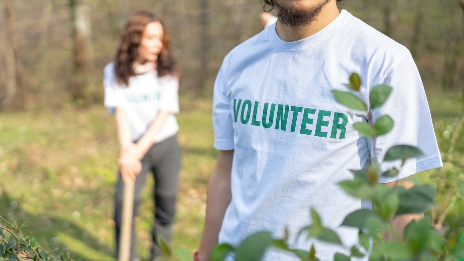 Ein Mann und eine Frau stehen auf einer Wiese vor einem Wald. Der Mann trägt ein T-Shirt auf dem ein Schriftzug mit dem Wort „Volunteer” (Freiwilliger) steht