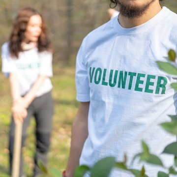 Ein Mann und eine Frau stehen auf einer Wiese vor einem Wald. Der Mann trägt ein T-Shirt auf dem ein Schriftzug mit dem Wort „Volunteer” (Freiwilliger) steht