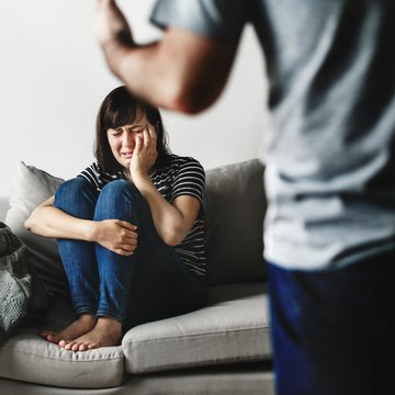 Das Foto zeigt eine junge Frau, die auf einem Sofa kauert und eine Hand vor ihr Gesicht hält. Vor ihr steht ein Mann. Diesen sieht man nur leicht angeschnitten von hinten. Er gestikuliert wild.