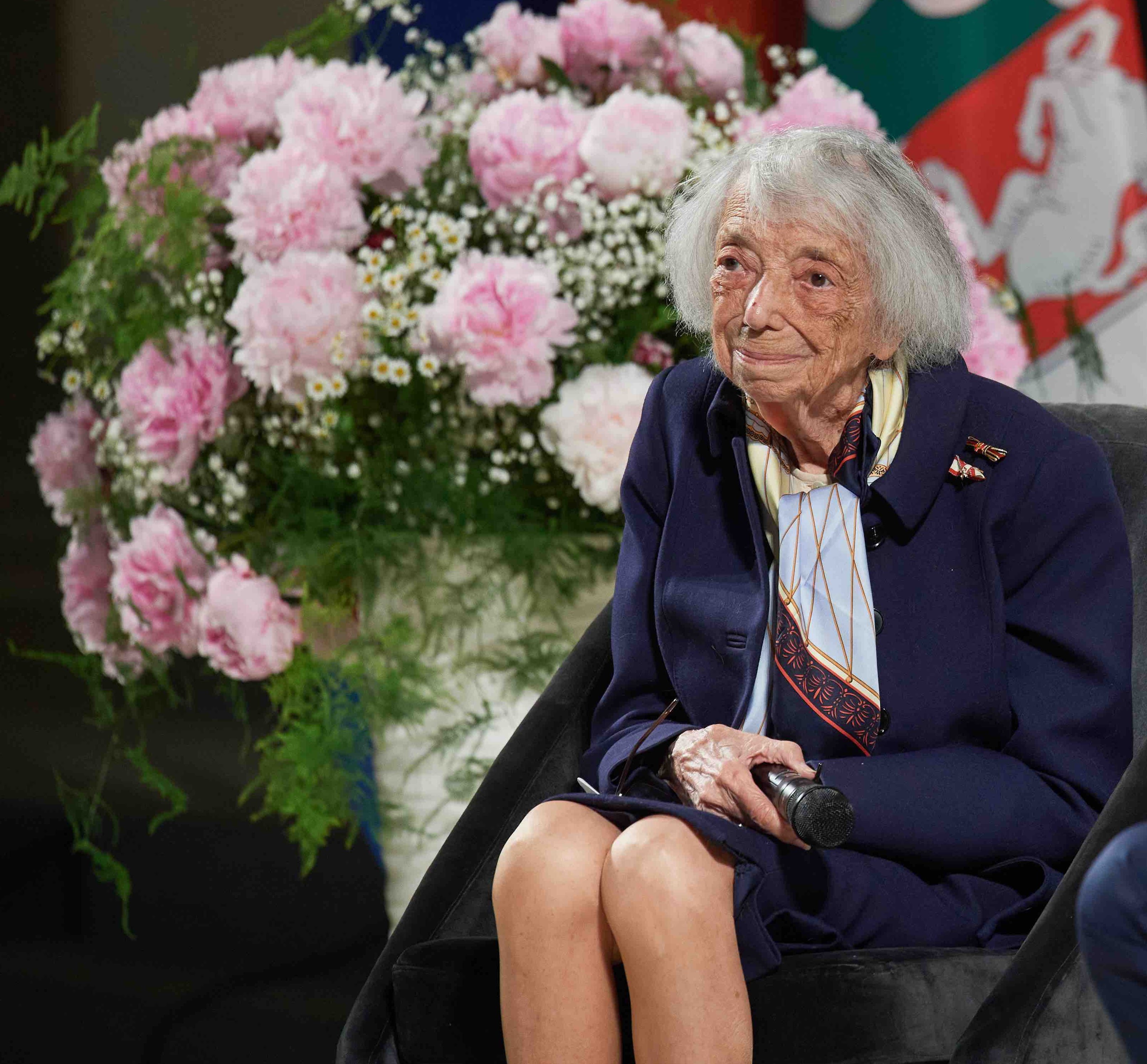 Die Holocaust-Überlebende Margot Friedländer sitzt auf einem Stuhl, links neben ihr ist ein großes Blumengesteck zu sehen.
