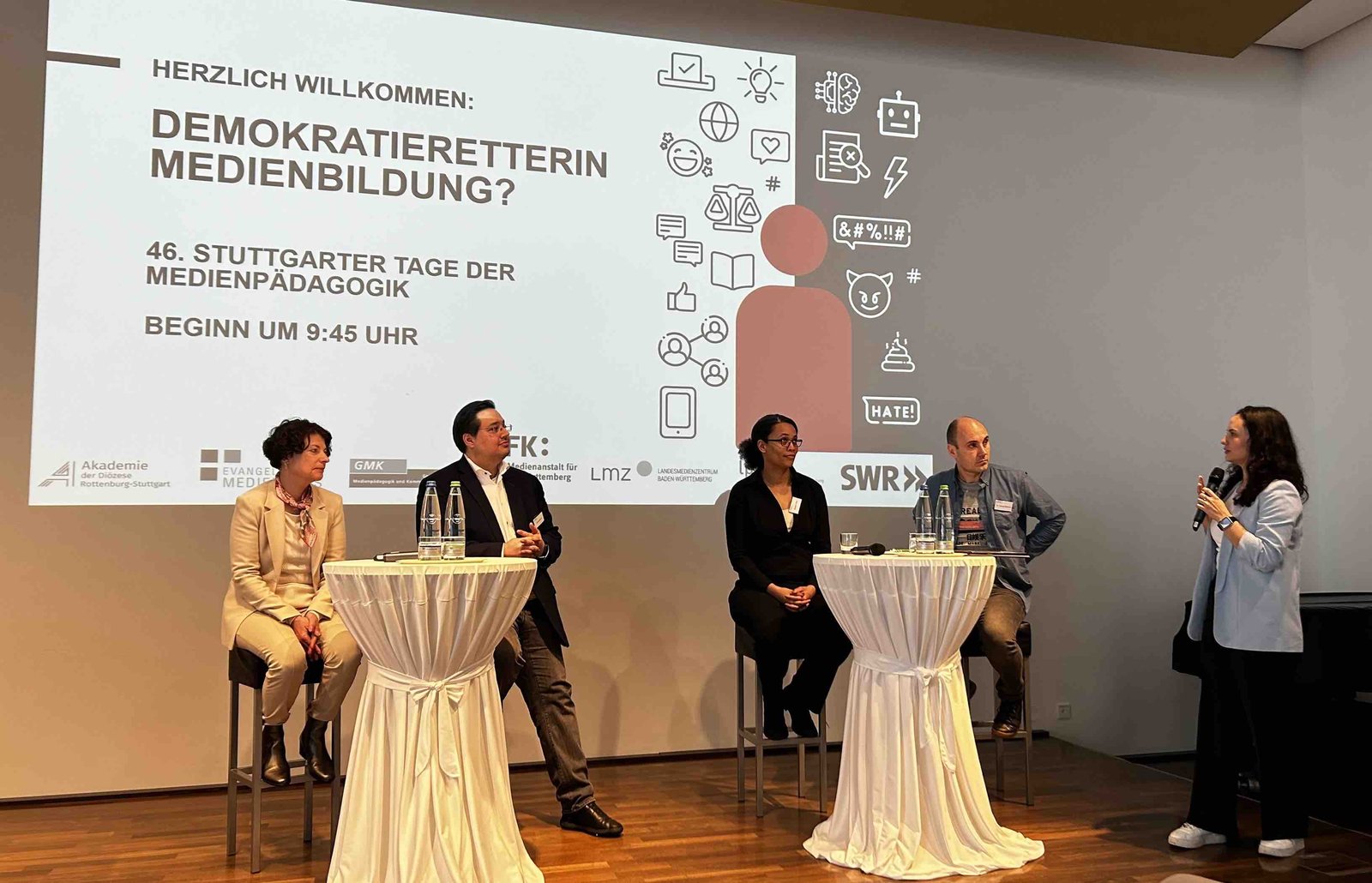 Drei Frauen und zwei Männer sind auf einer Bühne. Eine Frau hält ein Mikrofon in der Hand.