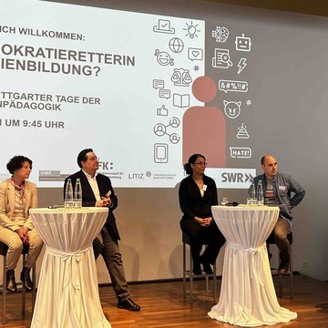 Drei Frauen und zwei Männer sind auf einer Bühne. Eine Frau hält ein Mikrofon in der Hand.