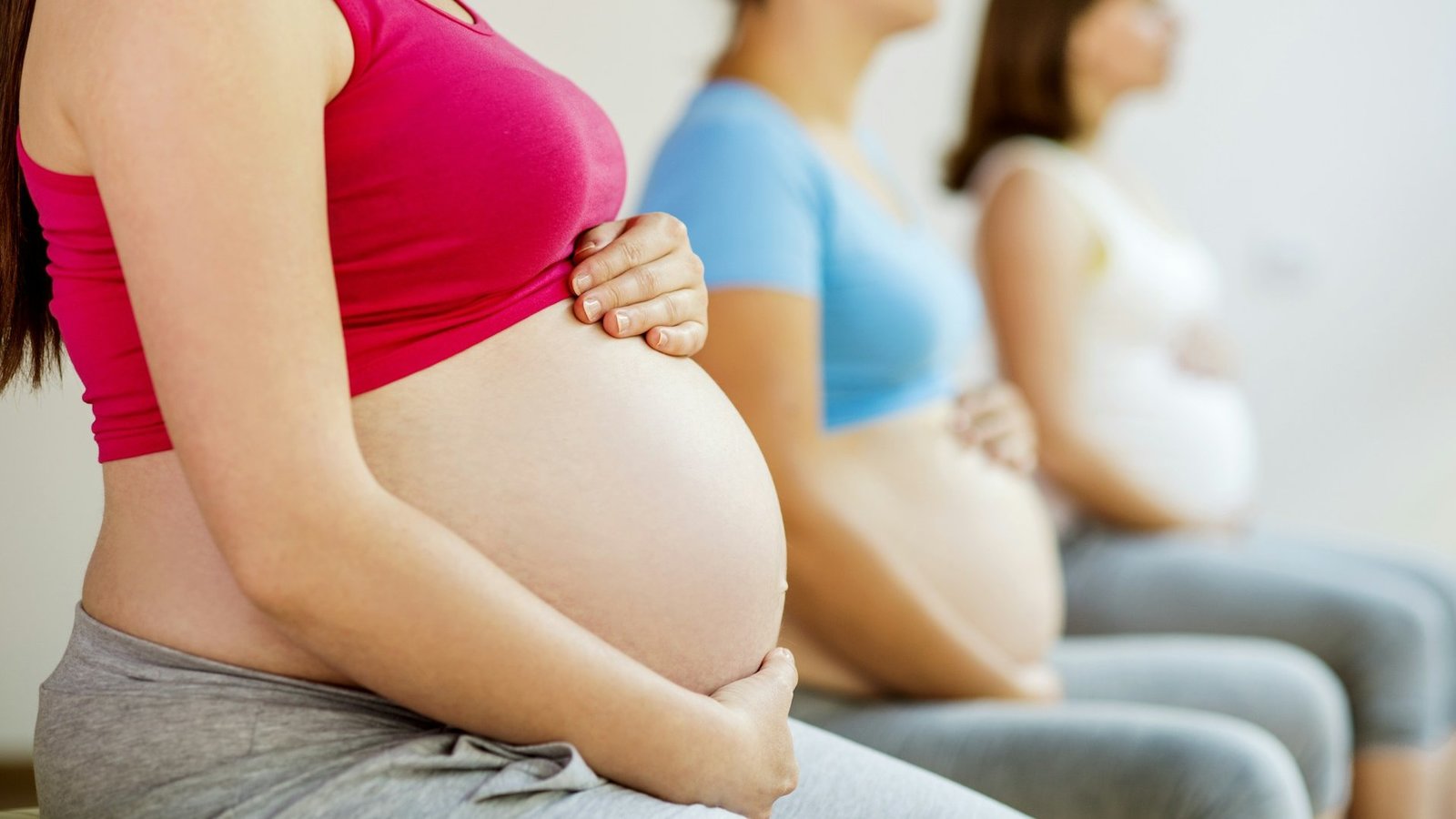 drei Frauen sitzen nebeneinander. Alle drei sind schwanger und halten ihre Hände um ihren jeweiligen Bauch.