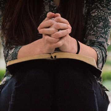 Man sieht eine Frau, die sich hinkniet und ihre zum Gebet gefalteten Hände auf ein offenes Buch abstützt. Der Kopf ist abgeschnitten. Die Frau ist nur vom Oberkörper abwärts bis zu den Knien zu sehen