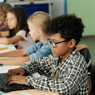 Seitenansicht von vier Kindern, die nebeneinander sitzen und jeweils einen Laptop vor sich auf einem Tisch stehen haben. 