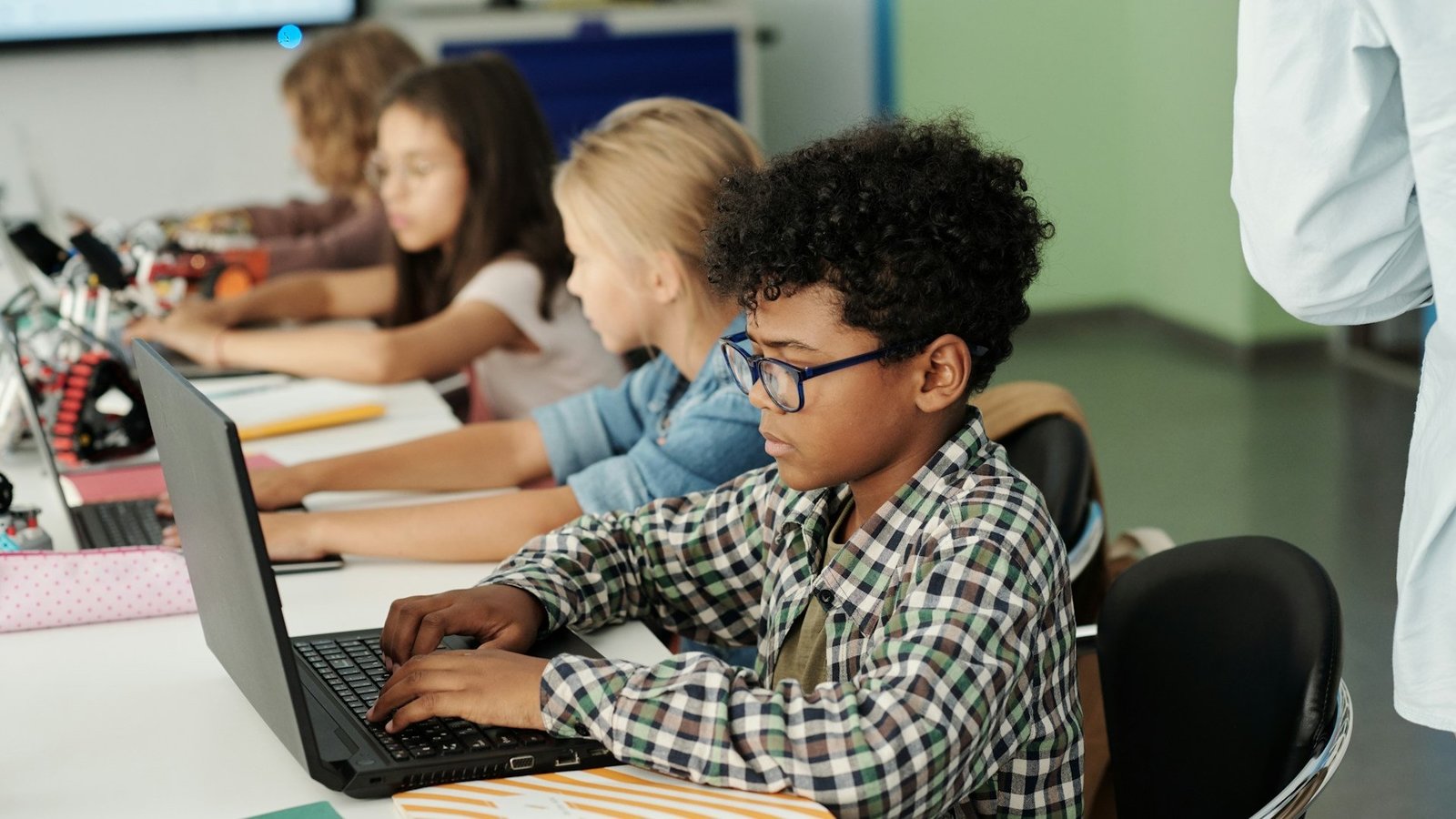 Seitenansicht von vier Kindern, die nebeneinander sitzen und jeweils einen Laptop vor sich auf einem Tisch stehen haben. 