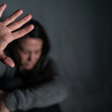 Eine Frau sitzt auf dem Boden, sie hat den Kopf nach unten gesenkt. Ihre rechte Hand habt sie abwehrend nach oben und zeigt ihre Handfläche.