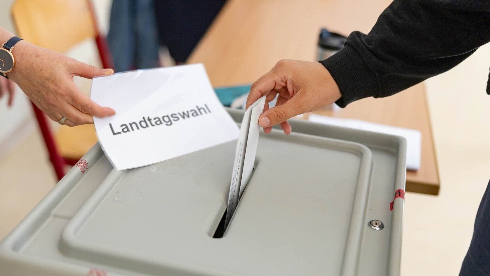 Eine Wahlurne mit einem Schlitz oben: Eine Person wirft ihren Wahlzettel in den Schlitz. Eine andere Person hält ein Schild mit der Aufschrift „Landtagswahl” in der Hand.