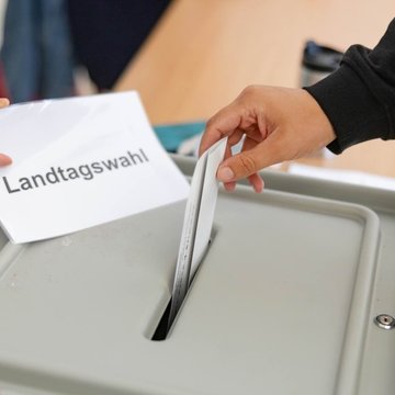 Eine Wahlurne mit einem Schlitz oben: Eine Person wirft ihren Wahlzettel in den Schlitz. Eine andere Person hält ein Schild mit der Aufschrift „Landtagswahl” in der Hand.