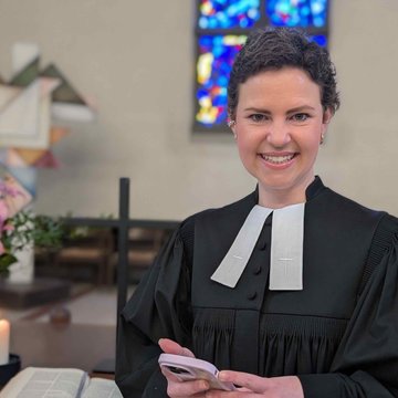 Sarah Schindler hat kurze braune Haare, sie trägt einen Talar mit weißen Beffchen und hält ein Handy in den Händen vor sich. Die junge Frau steht im Altarraum einer Kirche. Im Hintergrund sind Buntglasfenster zu sehen, eine Bibel, brennende Kerzen und ein Kreuz.