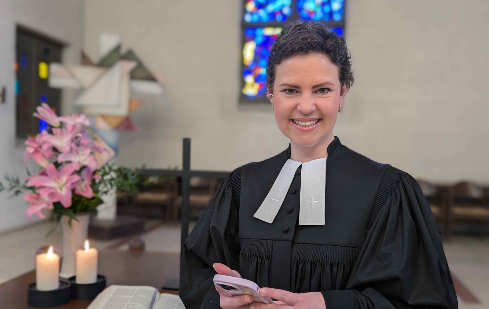 Sarah Schindler hat kurze braune Haare, sie trägt einen Talar mit weißen Beffchen und hält ein Handy in den Händen vor sich. Die junge Frau steht im Altarraum einer Kirche. Im Hintergrund sind Buntglasfenster zu sehen, eine Bibel, brennende Kerzen und ein Kreuz.