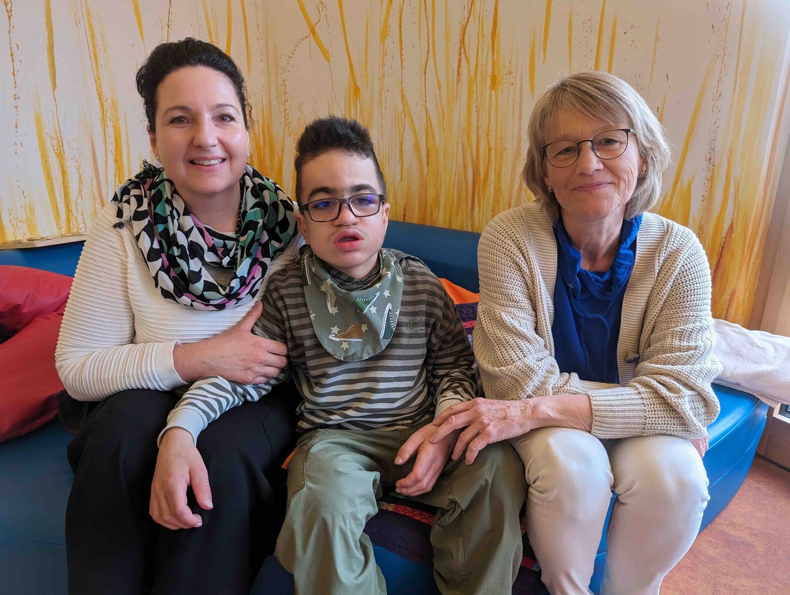 Sabine Beller und ihr 14-jähriger Sohn Silas sitzen gemeinsam mit der Gründerin Karin Eckstein auf einem blauen Sofa. Silas trägt eine Brille.