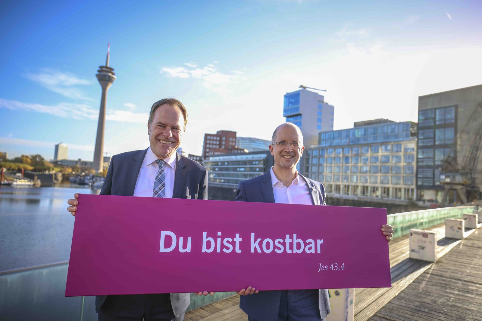 Von links: Der Düsseldorfer Oberbürgermeister Stephan Keller (CDU) und der Präses der Evangelischen Kirche im Rheinland Thorsten Latzel. Die beiden Männer halten ein magentafarbenes Schild in der Hand auf dem steht „Du bist kostbar Jes 43,4”. Die beiden Männer stehen auf einer Brücke im Hintergrund ist die Skyline von Düsseldorf zu sehen mit einem großen Turm.