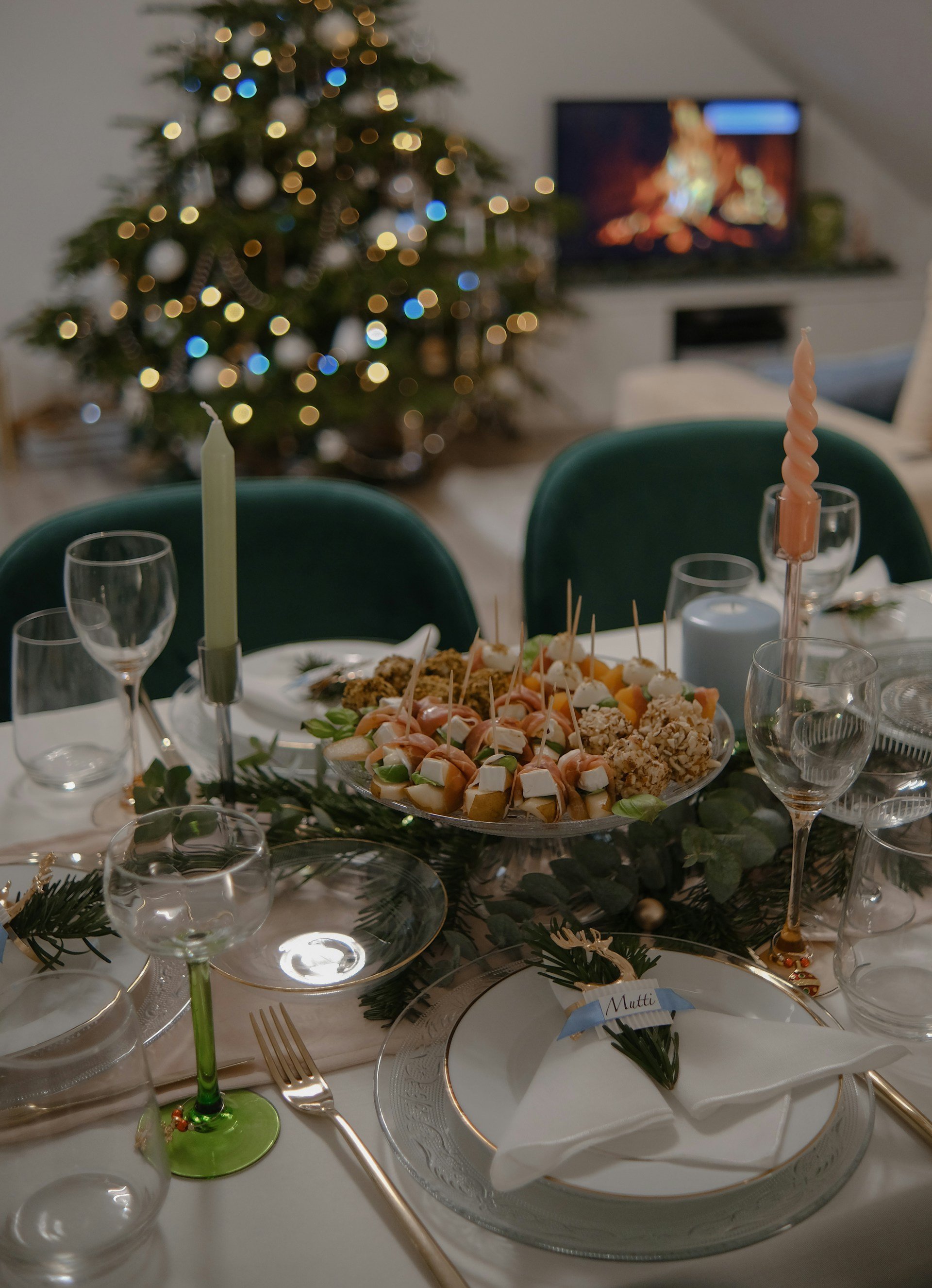 Es ist ein Wohnzimmer mit einem Weihnachtsbaum zu sehen, vor dem ein weihnachtlich gedeckter Esstisch und eine Vorspeise steht.
